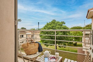 Un balcón con una cesta de mimbre, una tetera blanca y una taza sobre una mesa, con vistas a un paisaje verde y edificios.