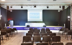 Un salón de conferencias con un escenario, sillas y una pantalla que muestra "Odaly's VACANCES".