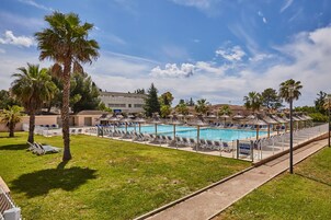 Un área de piscina con sillones de descanso y sombrillas, rodeada de palmeras y un cielo despejado.