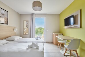A hotel room with two beds, a TV mounted on the wall, a desk with chairs, and a balcony with a view of greenery.