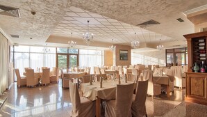 A spacious dining area with multiple tables and chairs, large windows, chandeliers, and a marble ceiling. - Park hotel Arbanasi (Veliko Tarnovo)