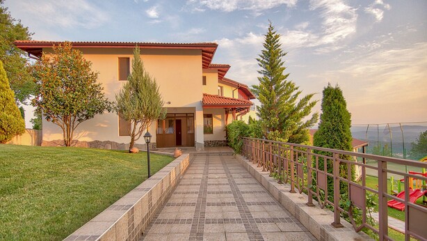 A two-story house with a tiled pathway leading to the entrance, surrounded by greenery and a view of the landscape. - Park hotel Arbanasi (Veliko Tarnovo)