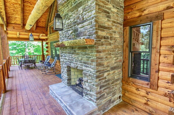 A wooden cabin with a stone fireplace, a covered porch with chairs, and a window overlooking greenery. - Whispering Trees Lodge (South Bloomingville)