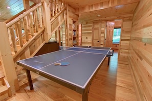 A ping pong table in a wooden room with a staircase and bookshelves. - Whispering Trees Lodge (South Bloomingville)