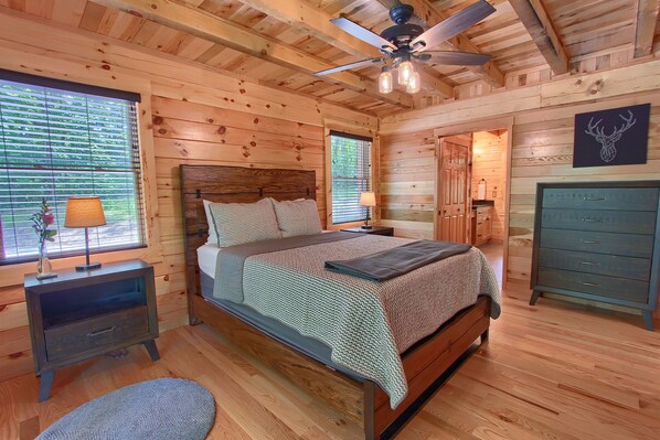 A wooden cabin bedroom with a bed, bedside tables, a dresser, a ceiling fan, and a window with blinds.