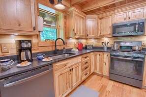 A kitchen with wooden cabinets, stainless steel appliances, a sink, and a window with a view of trees. - Whispering Trees Lodge (South Bloomingville)