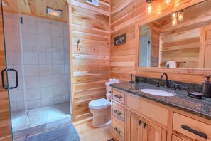 Een houten badkamer met een douchecabine van glas, een toilet, een wastafel met een granieten blad en houten kastjes.