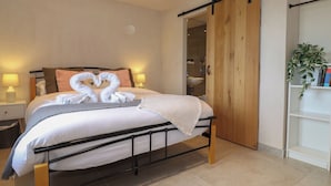 A neatly made bed with a heart-shaped towel arrangement, a bedside table with a lamp, and a sliding wooden door leading to a bathroom. - Meadow Syke Barn (Penrith)