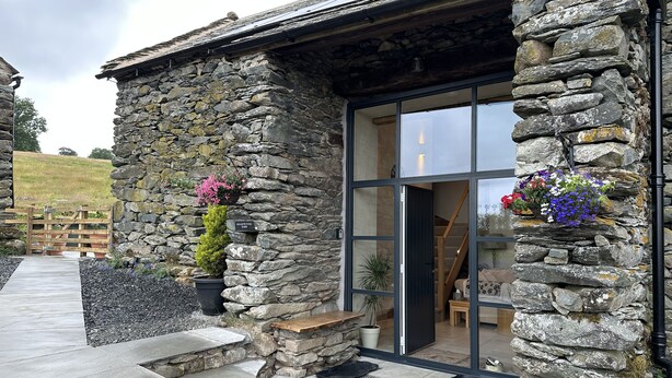 A stone building with a glass door, a wooden bench, and a stone wall with flower pots. - Meadow Syke Barn (Penrith)