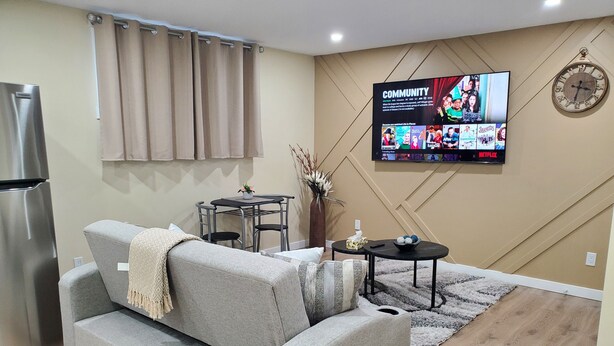 Un salon avec un canapé gris, une petite table, un téléviseur fixé au mur et une horloge.
