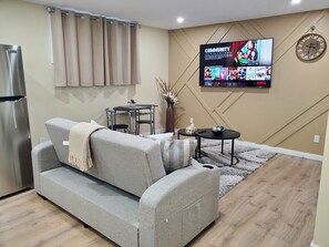 A living room with a grey sofa, a small table, a TV mounted on the wall, and a clock. - Paradise in the Meadows (Saskatoon)
