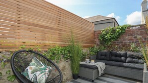 A patio with a black wicker chair, a grey sofa, and a wooden slatted wall. - 15 Rosemary Lane (Beaumaris)