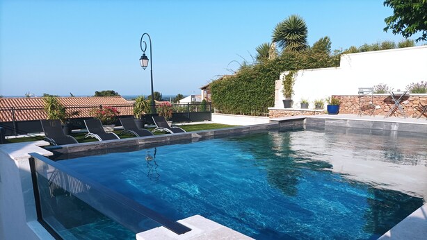 Une piscine à l’eau d’un bleu limpide, entourée d’un mur blanc et d’un mur en pierre. On y trouve des chaises longues et une plante en pot près de la piscine.