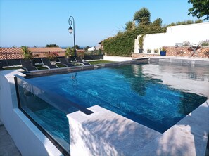 Ein Swimmingpool mit klarem, blauem Wasser, umgeben von einer weißen Mauer und einer Steinmauer. In der Nähe des Pools befinden sich Liegestühle und ein Blumentopf.