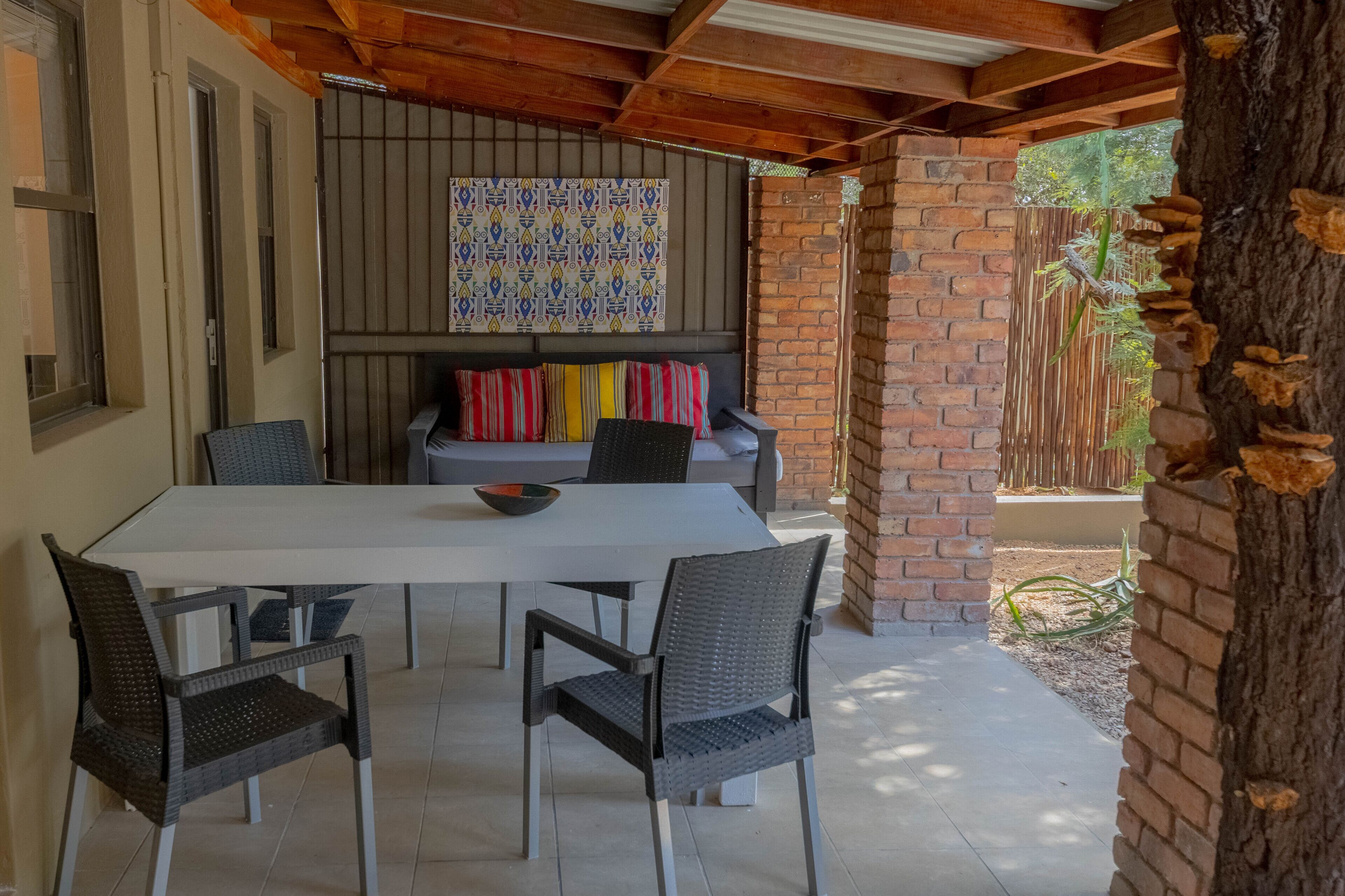 Une terrasse extérieure avec une table blanche et quatre chaises en osier, une décoration murale à motifs au-dessus de la table, et un mur de briques agrémenté d’une treille en bois.