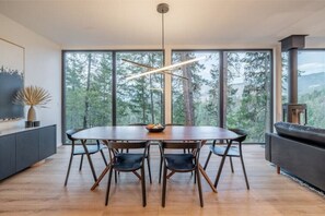 Enjoy meals in this spacious dining area with a large round table and six comfortable chairs, surrounded by expansive windows offering a view of the forest.