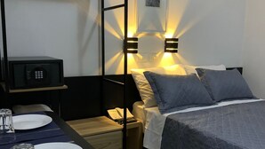 A compact hotel room with a bed, bedside table, and a small desk with a microwave, plates, and glasses.
