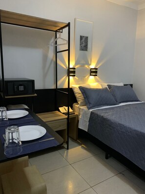 A compact hotel room with a bed, bedside table, and a small desk with a microwave, plates, and glasses. - Lory Studio Hotel (São Paulo)