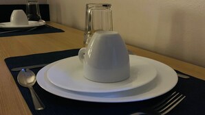 Uma mesa de jantar com uma chávena branca, um jarro de vidro, uma colher e um garfo sobre um guardanapo escuro.