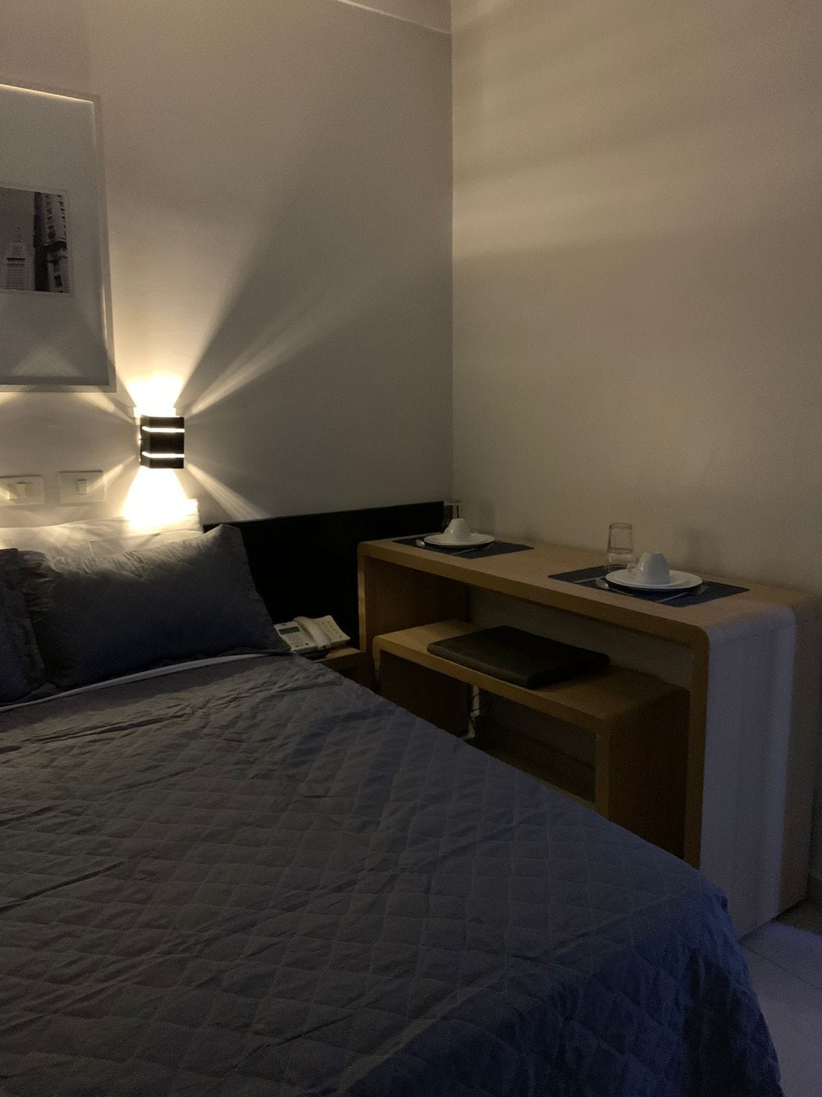 A hotel room with a bed, bedside table, and a wall-mounted light fixture.