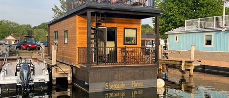 A wooden floating house with a balcony, docked in calm water, surrounded by greenery and other boats.