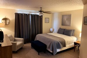 Ein Hotelzimmer mit einem Bett, einem Sessel, einem Schreibtisch, einem Deckenventilator und einem Fenster mit Vorhängen.