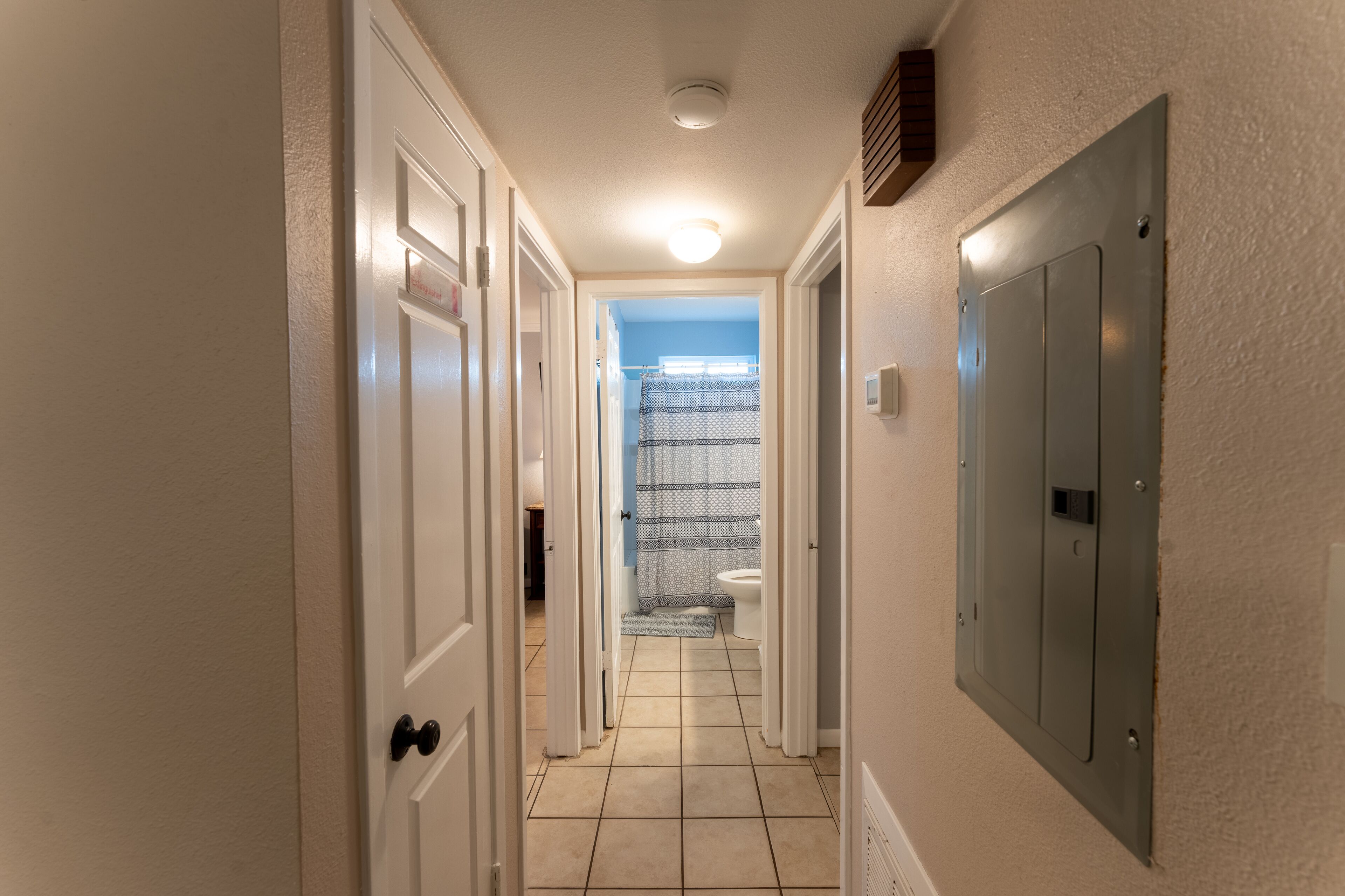 Un couloir étroit avec des portes blanches, un sol carrelé et une salle de bain visible, séparée par un rideau de douche.