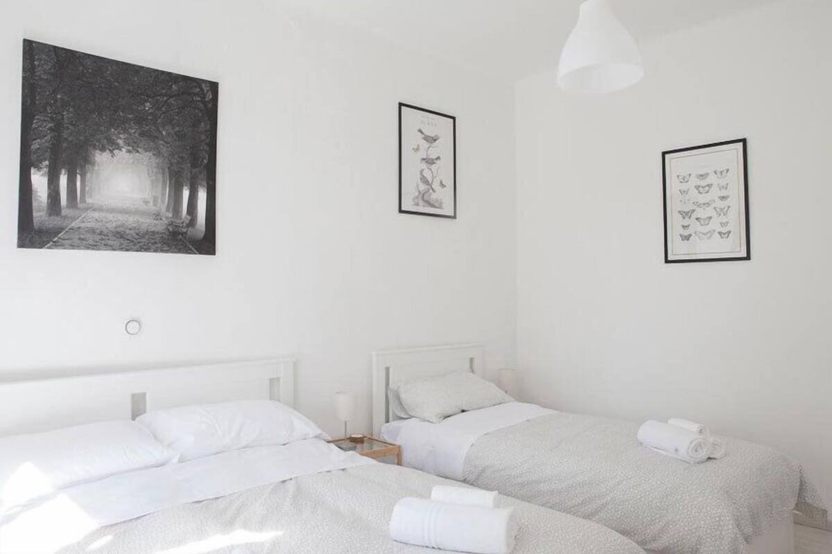 A minimalist hotel room with two beds, a large framed picture of a forest path, and two smaller framed pictures on the walls.