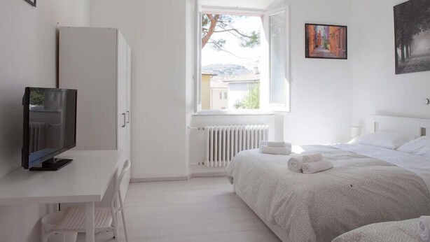 A hotel room with a bed, a desk with a computer, a radiator, and a window with a view of buildings. - Cleo Giglio  (Ancona)