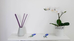 A white shelf with a white vase containing purple reed sticks, a white orchid, and two keychains. - Cleo Giglio  (Ancona)