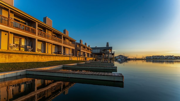 A waterfront hotel with a dock extending into calm water, featuring multiple balconies and a serene sunset view. - Waterfront Condo: Stunning Sunsets, Fishing, Patio, & 4 minutes from the beach!  (Corpus Christi)