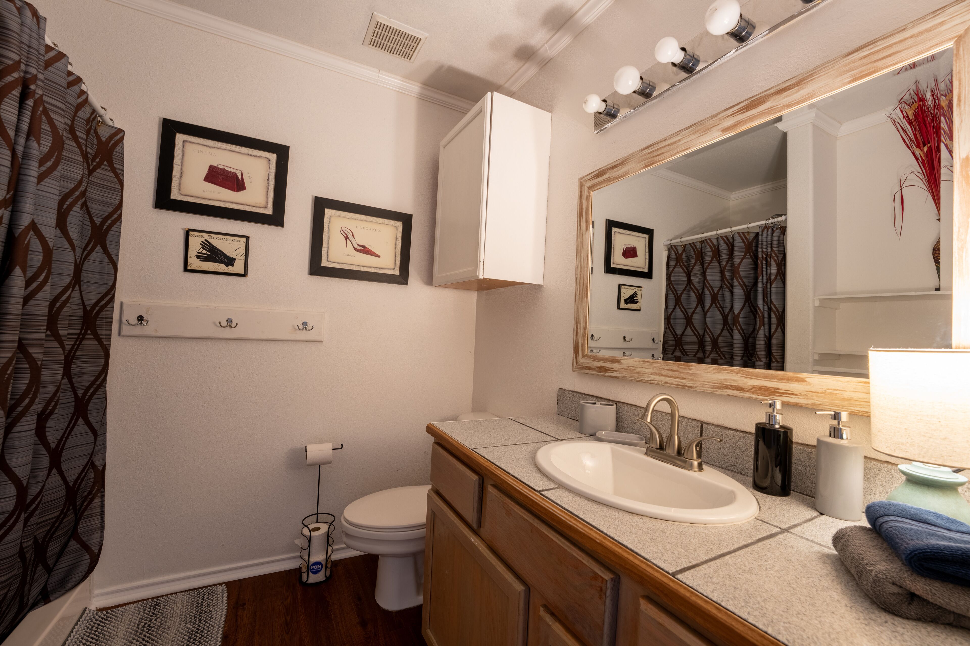Une salle de bain avec un lavabo, un meuble-lavabo et un miroir blancs. On y trouve des tableaux encadrés au mur, des toilettes et un rideau de douche.