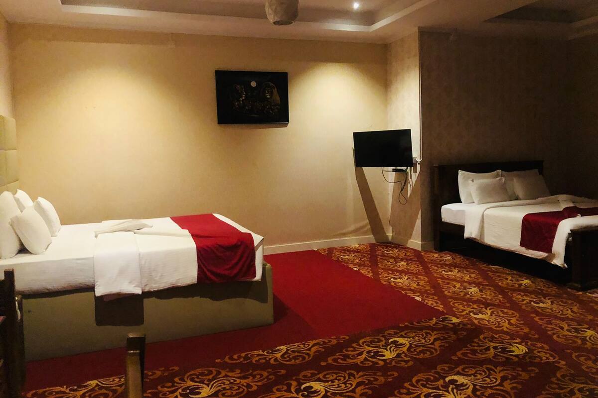 Ein Hotelzimmer mit zwei Betten, einem roten Teppich, einem Gemälde an der Wand und einem an der Wand befestigten Fernseher.