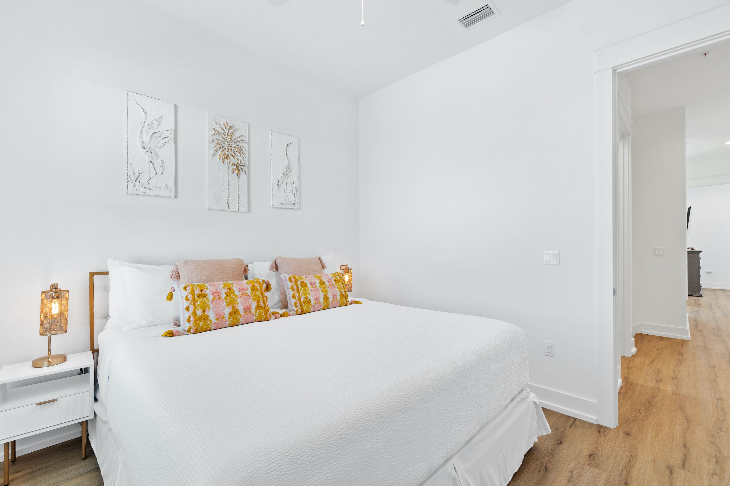 Un dormitorio con una cama grande, paredes blancas y suelo de madera. Hay tres cuadros en la pared y una mesita de noche con una lámpara.
