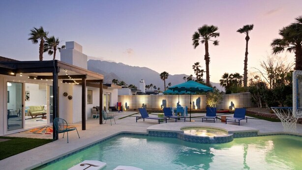 A swimming pool with lounge chairs and a covered area, surrounded by palm trees and a mountain backdrop.