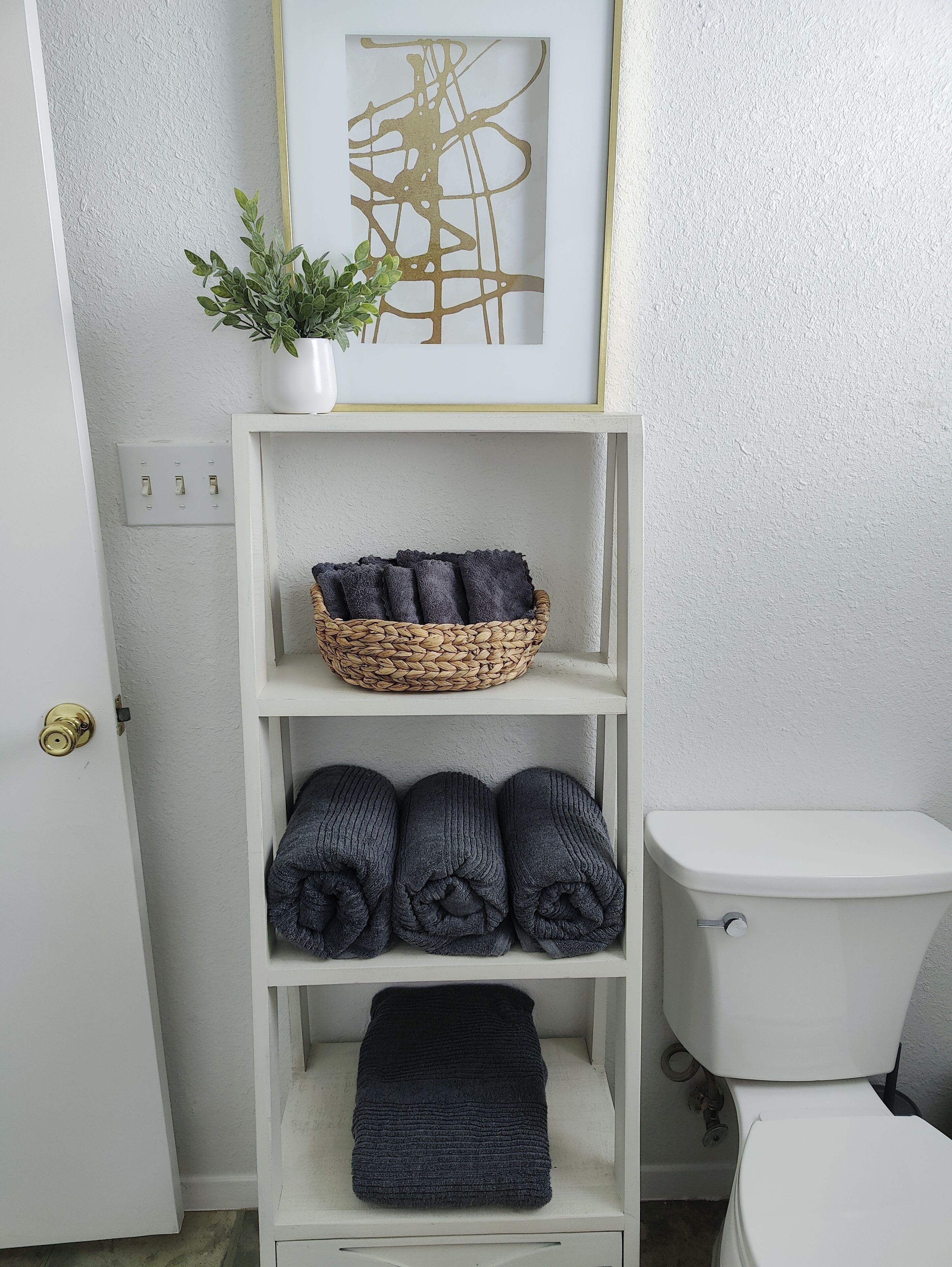 A bathroom with a white toilet, a shelf with folded towels, a woven basket, and a framed abstract artwork.