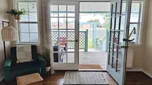 Una habitación con un sofá verde, un banco de madera, un macramé colgante, una lámpara de mimbre y vistas a una terraza exterior a través de un ventanal.