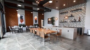 A modern dining area with a long wooden table and chairs, a kitchen area with a tiled backsplash, and a stone tile floor.