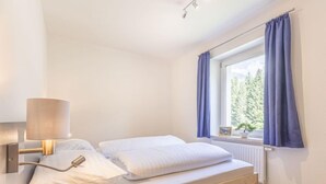 Un dormitorio con cama, lámpara de noche, ventana con cortinas y vistas a árboles.