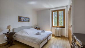 Un dormitorio con cama, mesita de noche, cómoda, ventana con vistas a zonas verdes y un radiador.