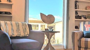 A living room with a sofa, a table with a decorative bowl, and a view of a building through a large window. - The Dolphin: Beach~Workspace~W/D incl~PetsOK (Long Beach)