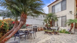 A patio area with outdoor furniture, a round table, and chairs. There are palm trees and a building with large windows in the background. - The Dolphin: Beach~Workspace~W/D incl~PetsOK (Long Beach)