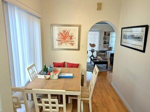 A dining area with a wooden table, chairs, and a red cushion. There is a framed artwork on the wall and a framed photo on the wall to the right.