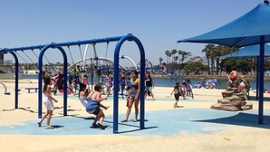 A playground with swings and climbing structures, surrounded by a sandy area and palm trees. - The Dolphin: Beach~Workspace~W/D incl~PetsOK (Long Beach)