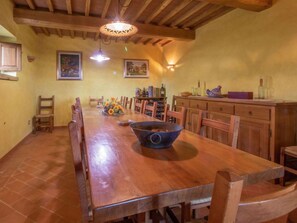 Ein Essbereich mit einem langen Holztisch, Stühlen und einer dekorativen Schale in der Mitte. Wandleuchten und ein Weinkühlschrank mit Flaschen sind ebenfalls vorhanden.