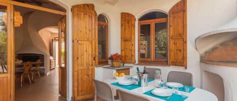 A dining area with a white table set for four, wooden shutters on the windows, and a stone oven in the background.