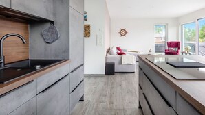 Une cuisine moderne avec des meubles gris, un plan de travail en bois et une cuisinière encastrée. Le salon est doté d’un canapé gris agrémenté d’un coussin rouge et d’une couverture blanche. On aperçoit une chaise rouge près de la fenêtre.
