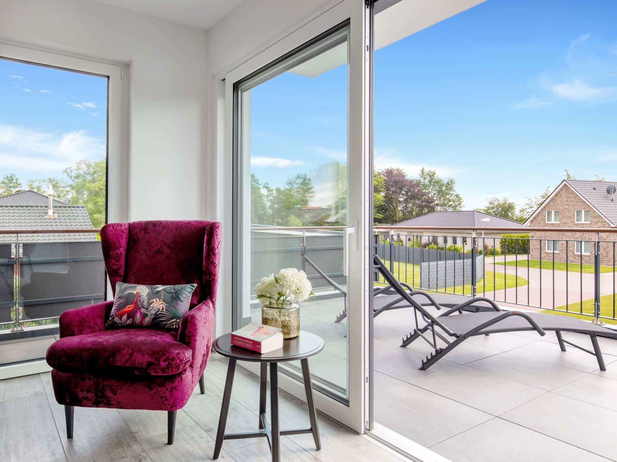 A balcony with a red velvet chair, a small table with a book and flowers, and a clear view of houses and greenery.
