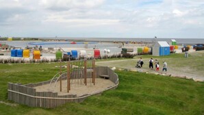 Une zone aménagée au bord de la plage, avec une structure de jeux en bois, un petit abri et des cabanes de plage aux couleurs vives.