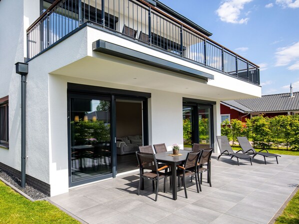 A modern two-story house with a balcony, a patio with a dining table and chairs, and a garden area.